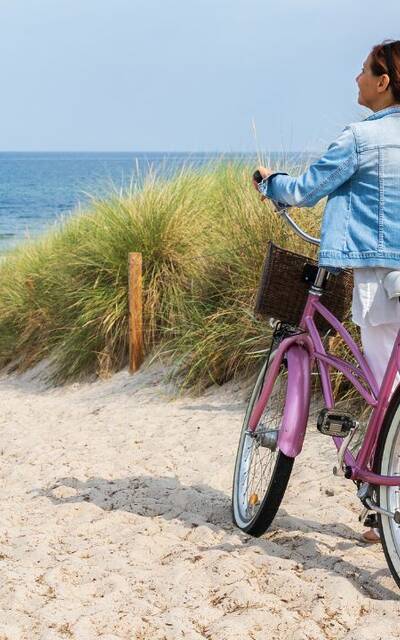 Photo d'une dame avec son vélo rose. Elle marche en direction de la plage dans un chemin de sable entouré de fougères.